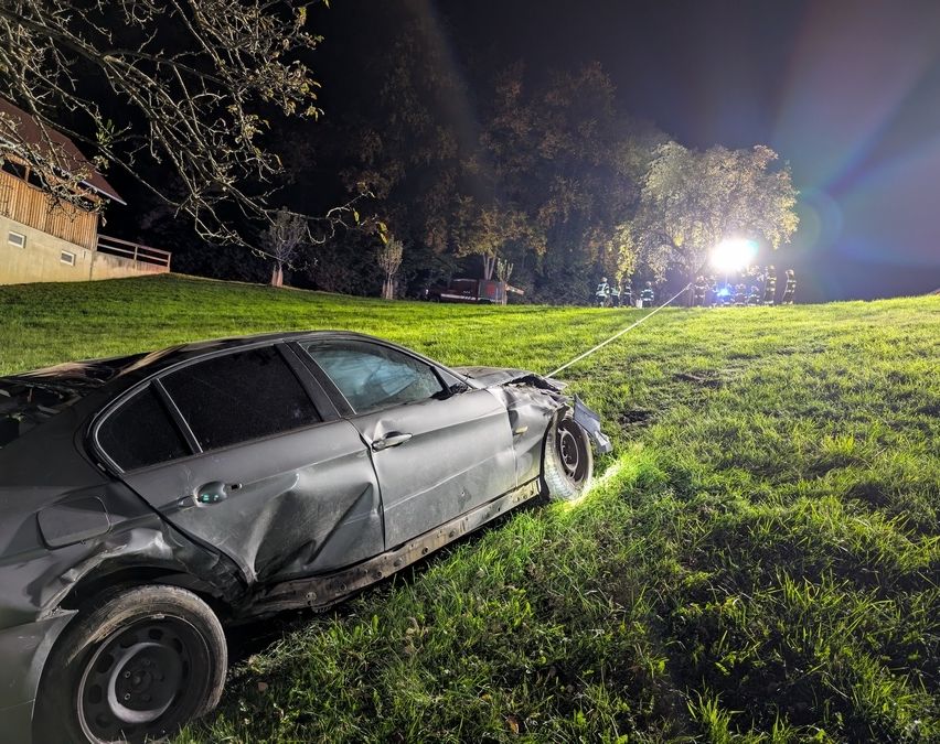 Ein beschädigtes Auto steht bei Nacht auf einem Rasenfeld, beleuchtet von einem Scheinwerfer. Mehrere Menschen, möglicherweise Rettungskräfte, stehen in der Nähe. Bäume und ein Gebäude sind im Hintergrund zu sehen.