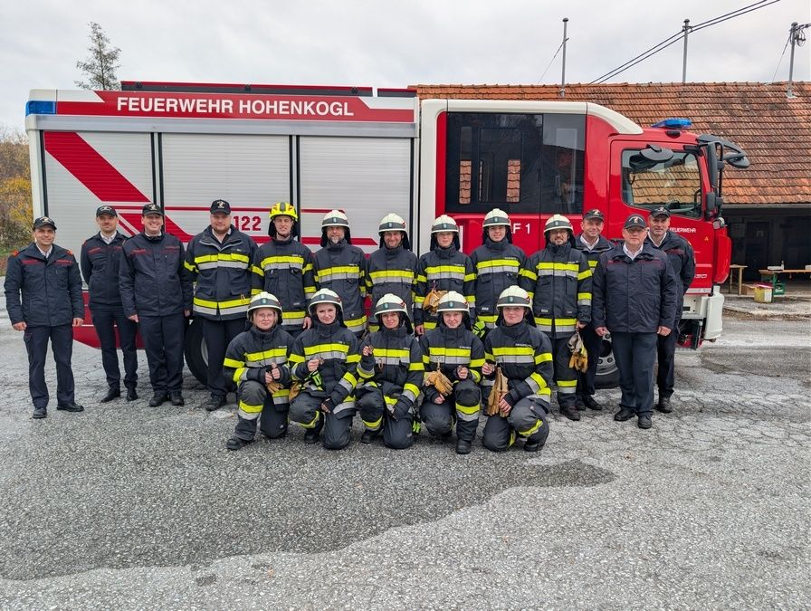 Eine Gruppe Feuerwehrleute in Uniform posiert vor einem Feuerwehrauto, mit Gebäuden und Bäumen im Hintergrund.