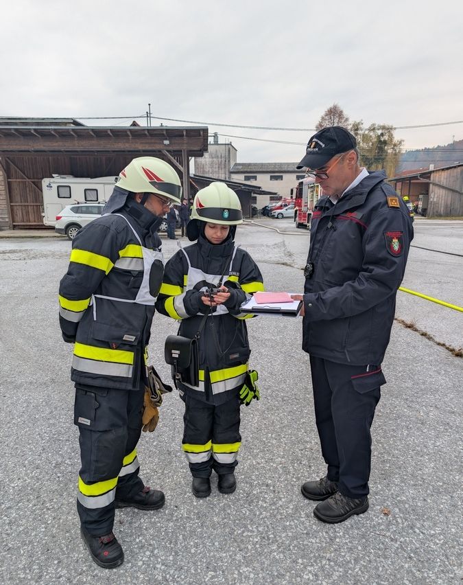 Drei Feuerwehrleute stehen auf einem Parkplatz. Zwei von ihnen tragen gelbe und schwarze Uniformen. Der dritte trägt einen Hut und hält eine Mappe. Hinter ihnen steht ein Gebäude mit einem geparkten Feuerwehrwagen vor der Tür.