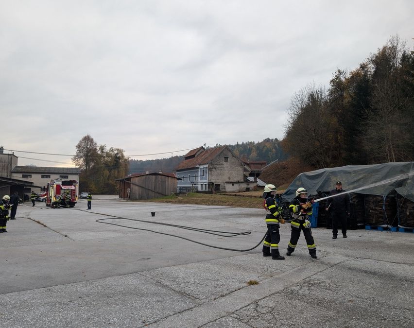 Feuerwehrleute üben mit einem Feuerwehrschlauch auf einem offenen Gelände, mit einem Feuerwehrauto und Gebäuden im Hintergrund.