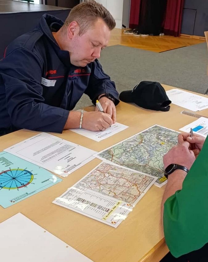 Ein Mann in einem dunkelblauen Jacken sitzt an einem Tisch und schreibt auf Papier, während vor ihm eine Karte ausgebreitet ist.