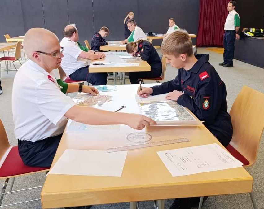 Eine Gruppe von Personen, möglicherweise Schüler, versammelt sich um einen Tisch in einer Klassenzimmerumgebung und beschäftigt sich mit einer Aktivität. Einige schreiben, andere schauen sich Karten an. Ein Mann in einem weißen Hemd zeigt auf eine Karte.