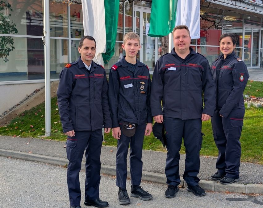 Vier uniformierte Personen, möglicherweise Feuerwehrleute, stehen vor einem Gebäude mit Fahnen im Hintergrund.