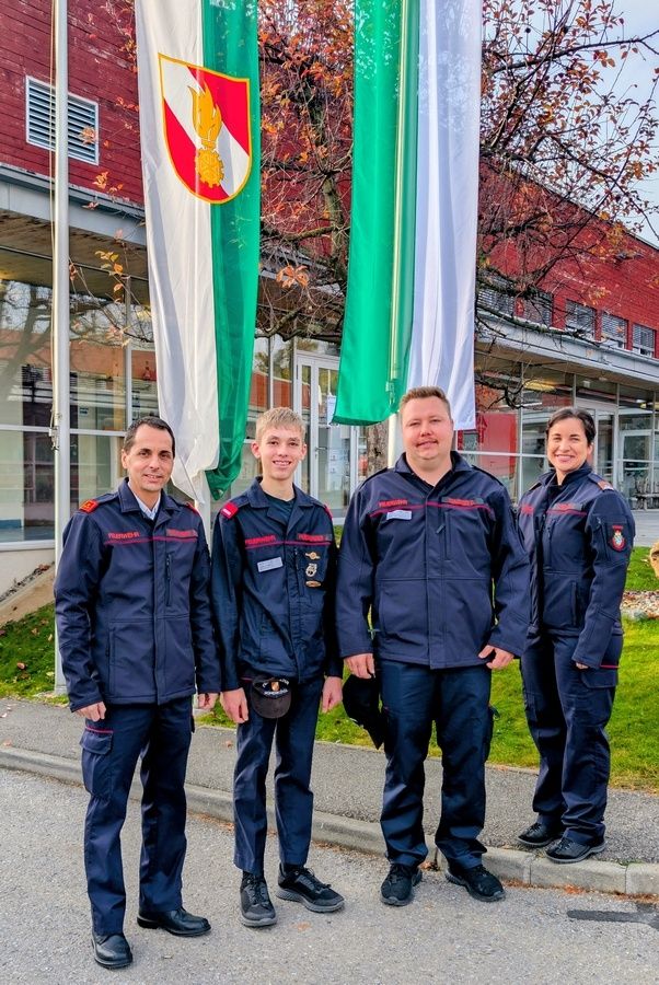 Fünf Menschen in Uniform stehen vor einem Gebäude mit Flaggen und lächeln für ein Foto. Hinter ihnen stehen rote und grüne Flaggen, und das Gebäude hat Glasfenster.