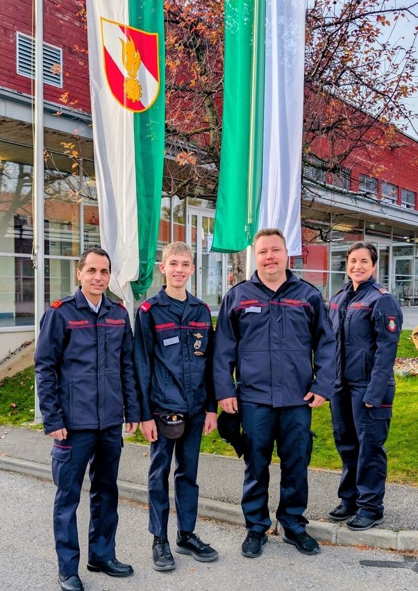 Fünf Menschen in Uniform stehen vor einem Gebäude mit Flaggen und lächeln für ein Foto. Hinter ihnen stehen rote und grüne Flaggen, und das Gebäude hat Glasfenster.