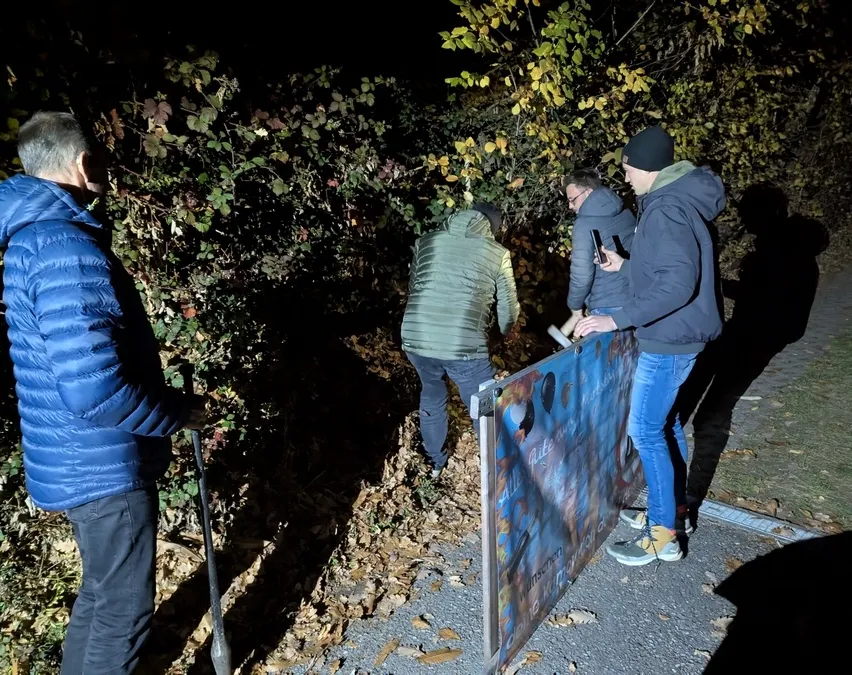 Drei Männer in warmer Kleidung sind nachts draußen, einer hält eine Schaufel. Sie scheinen etwas in den Büschen zu suchen.