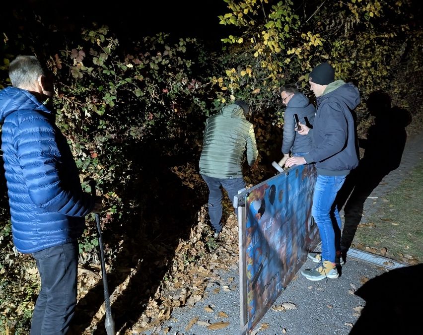Drei Männer in warmer Kleidung sind nachts draußen, einer hält eine Schaufel. Sie scheinen etwas in den Büschen zu suchen.