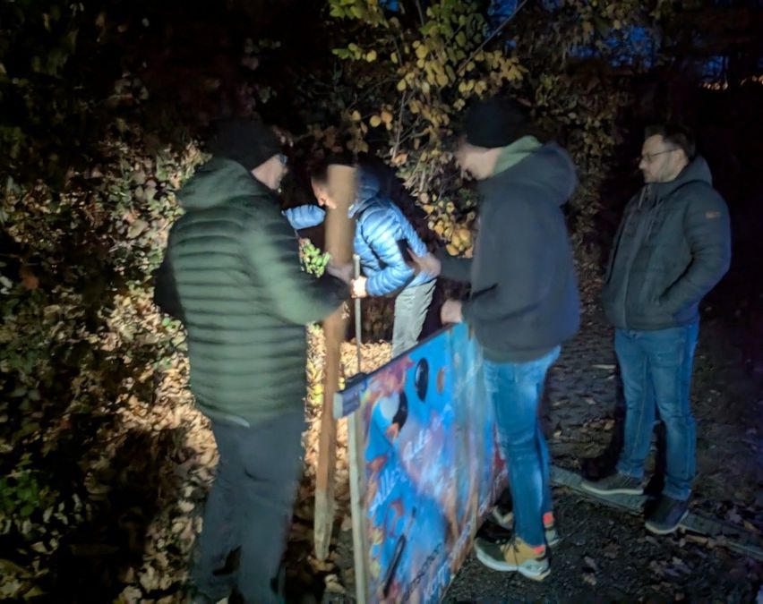 Vier Männer sind nachts draußen. Einer hält ein Banner mit blauer und schwarzer Kunst. Ein anderer beugt sich, um einen Holzpfosten zu platzieren. Sie stehen zwischen Bäumen und Blättern.