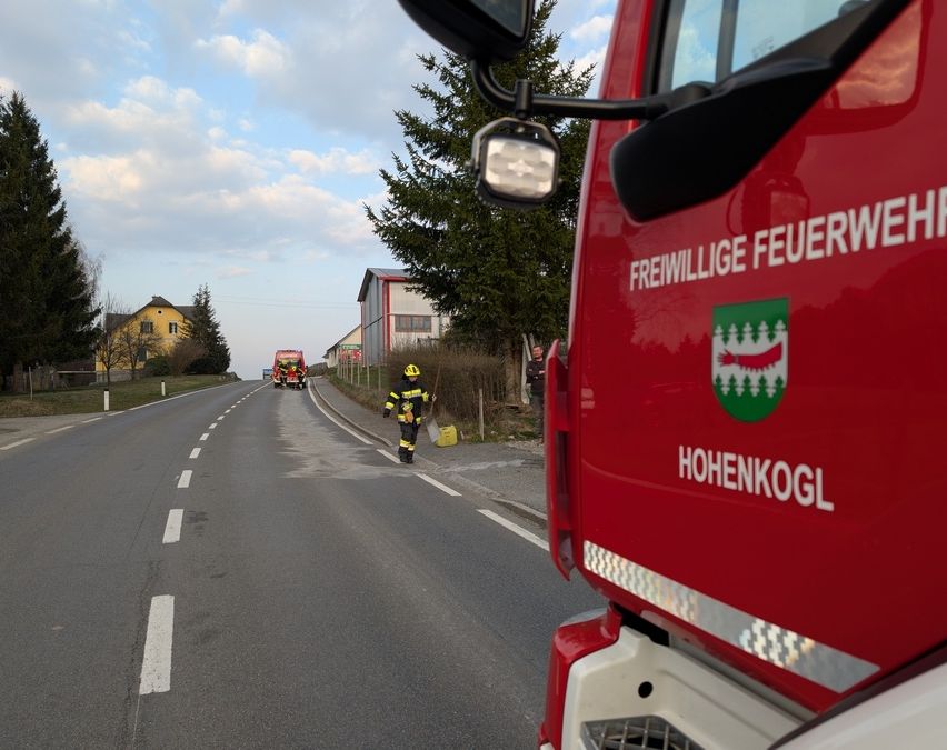 Ein freiwilliger Feuerwehrmann steht auf einer nassen Straße, hält einen Besen in der Hand, mit einem Feuerwehrwagen im Hintergrund und einem Gebäude in der Nähe.