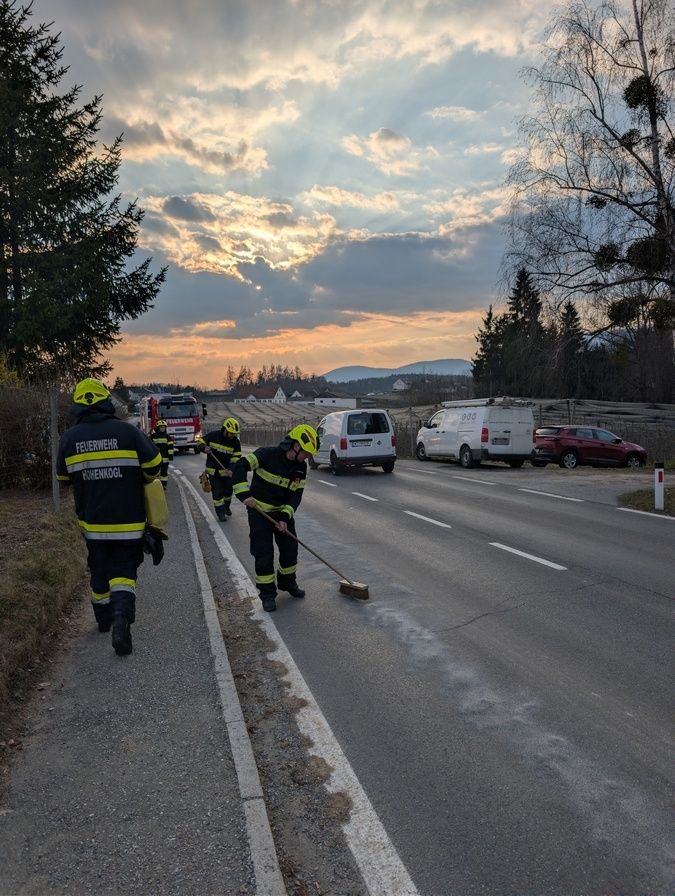 Feuerwehrleute in gelben Uniformen reinigen die Straße. Ein Feuerwehrwagen und Lieferwagen sind geparkt. Bäume und Berge im Hintergrund.