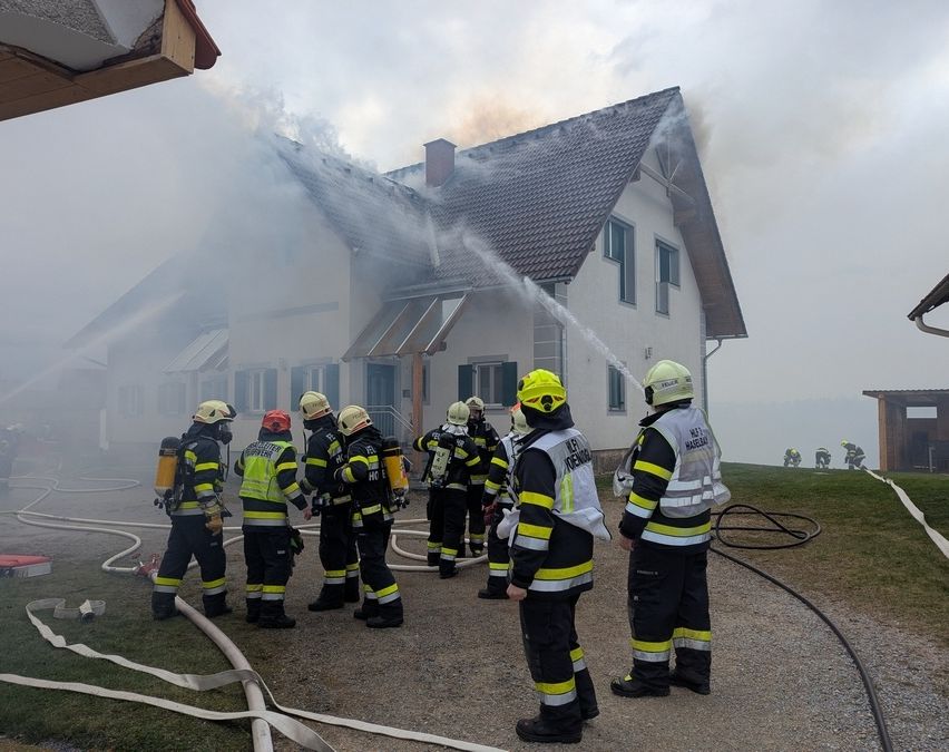 Feuerwehrleute stehen vor einem brennenden Haus, einer von ihnen spritzt Wasser, während andere in einer Reihe stehen.