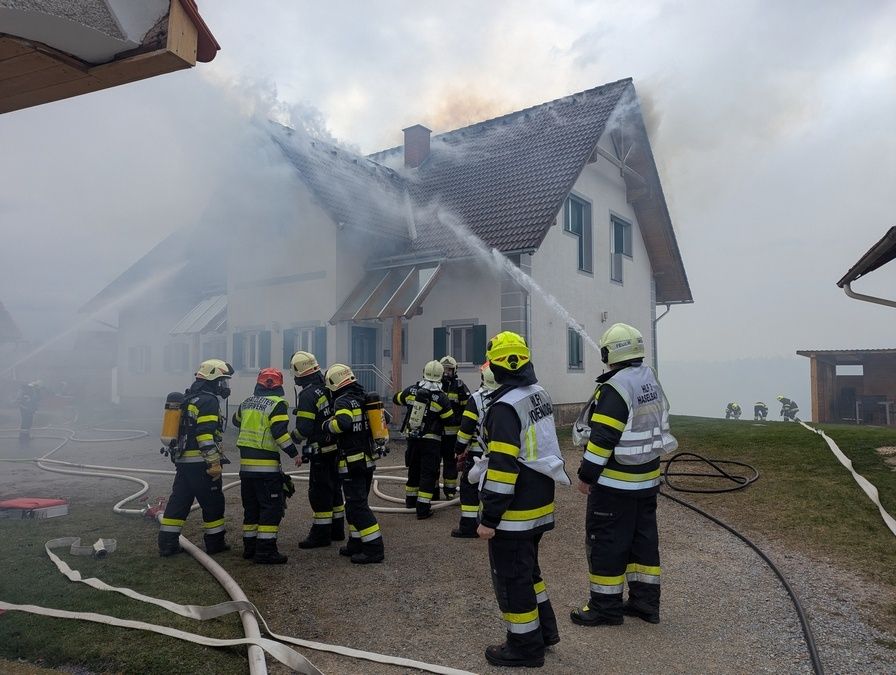 Feuerwehrleute stehen vor einem brennenden Haus, einer von ihnen spritzt Wasser, während andere in einer Reihe stehen.
