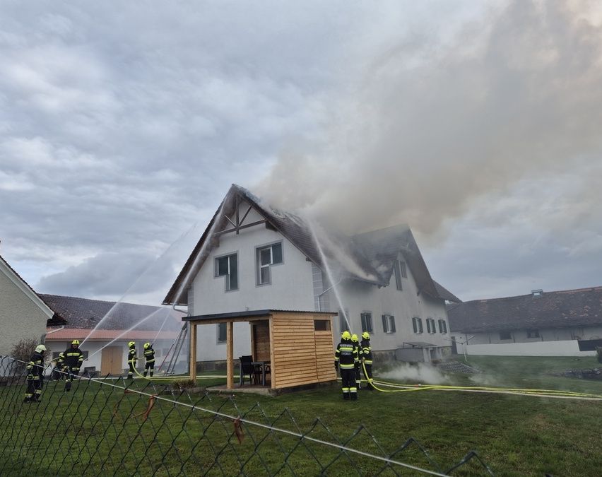 Feuerwehrleute löschen ein Feuer auf einem Haus. Rauch quillt aus dem Dach. Es gibt Feuerwehrleute am Boden und andere, die Wasser aus einer Feuerwehrspritze spritzen.