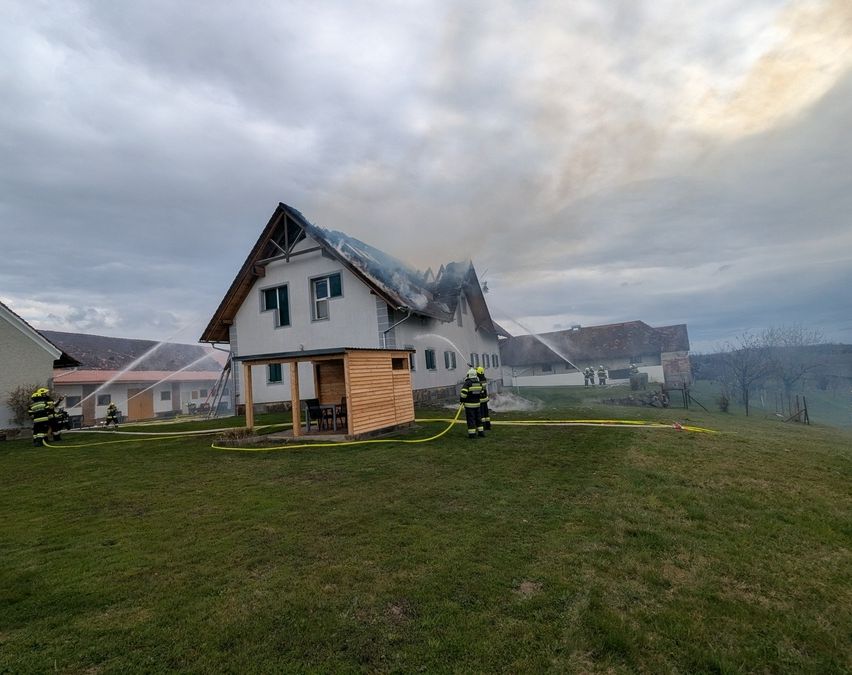 Feuerwehrleute sprühen Wasser auf ein brennendes Haus an einem bewölkten Tag. Das Haus ist von einem Grasfeld umgeben.