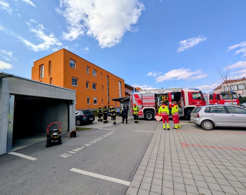 Eine Gruppe von Feuerwehrleuten in gelben Uniformen versammelt sich vor einem Gebäude mit orangefarbener Fassade, wahrscheinlich im Begriff, ein Feuer zu bekämpfen. Ein Feuerwehrwagen ist in der Nähe geparkt, seine Tür ist offen.