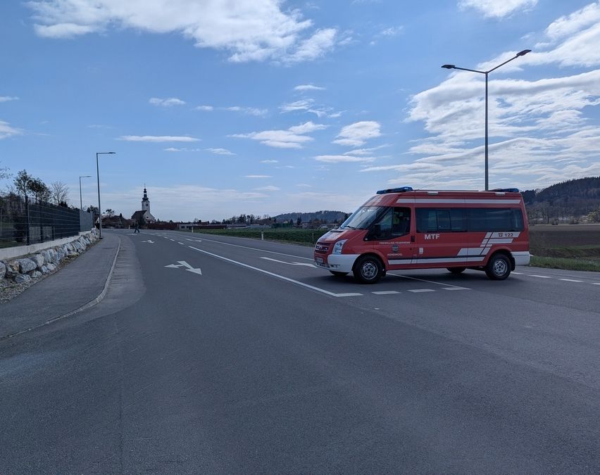 Ein Rettungsfahrzeug ist auf einer Straße geparkt, wobei Straßenlaternen und ein Gebäude in der Ferne unter einem teilweise bewölkten Himmel sichtbar sind.