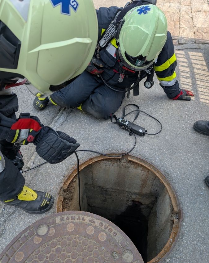 Zwei Feuerwehrleute in voller Ausrüstung überprüfen einen Kanaldeckel mit einem angeschlossenen Überwachungsgerät. Einer kniet und benutzt ein Gerät, während der andere steht und einen Draht hält.