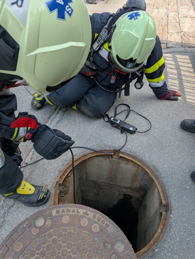 Zwei Feuerwehrleute in voller Ausrüstung überprüfen einen Kanaldeckel mit einem angeschlossenen Überwachungsgerät. Einer kniet und benutzt ein Gerät, während der andere steht und einen Draht hält.