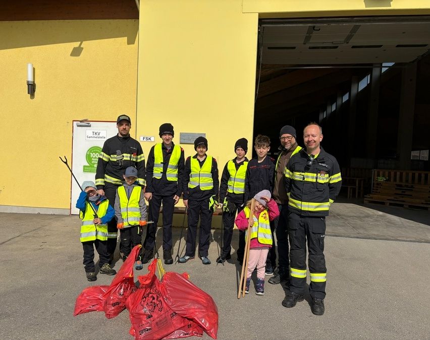 Eine Gruppe Feuerwehrleute, Kinder und Erwachsene stehen vor einem Gebäude, halten Stöcke und rote Müllsäcke und nehmen möglicherweise an einer Reinigungsaktion teil.