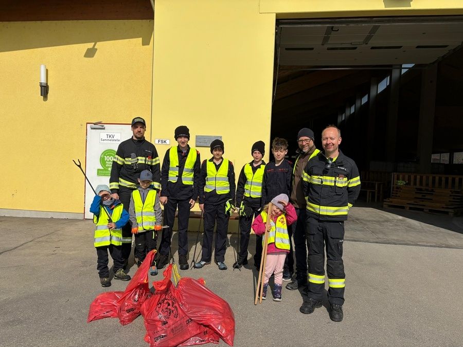 Eine Gruppe Feuerwehrleute, Kinder und Erwachsene stehen vor einem Gebäude, halten Stöcke und rote Müllsäcke und nehmen möglicherweise an einer Reinigungsaktion teil.