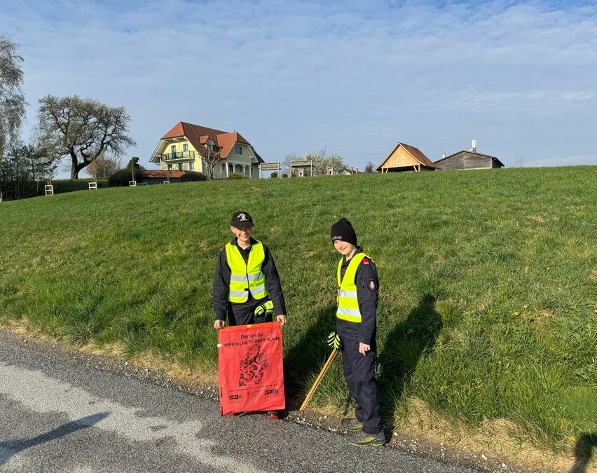 Zwei Personen in Warnwesten stehen neben einer Straße, eine hält ein rotes Banner und die andere einen Stock. Ein Haus und ein Baum sind im Hintergrund.
