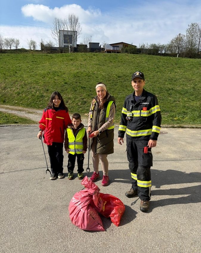 Drei Menschen in Uniform stehen in einem Park, halten Stöcke und lächeln. Zwei rote Taschen liegen auf dem Boden. Die Frau hält einen Stock und der Mann trägt eine Feuerwehruniform.