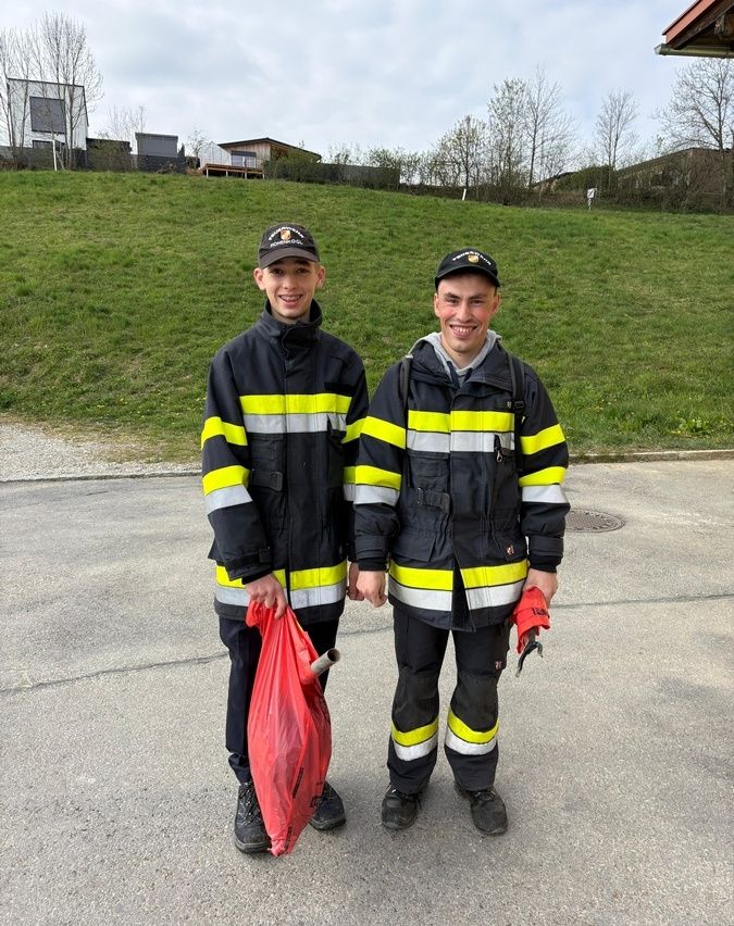 Zwei junge Männer in Feuerwehruniformen stehen auf einer Straße mit einem Hügel im Hintergrund. Sie lächeln und einer hält einen roten Beutel.