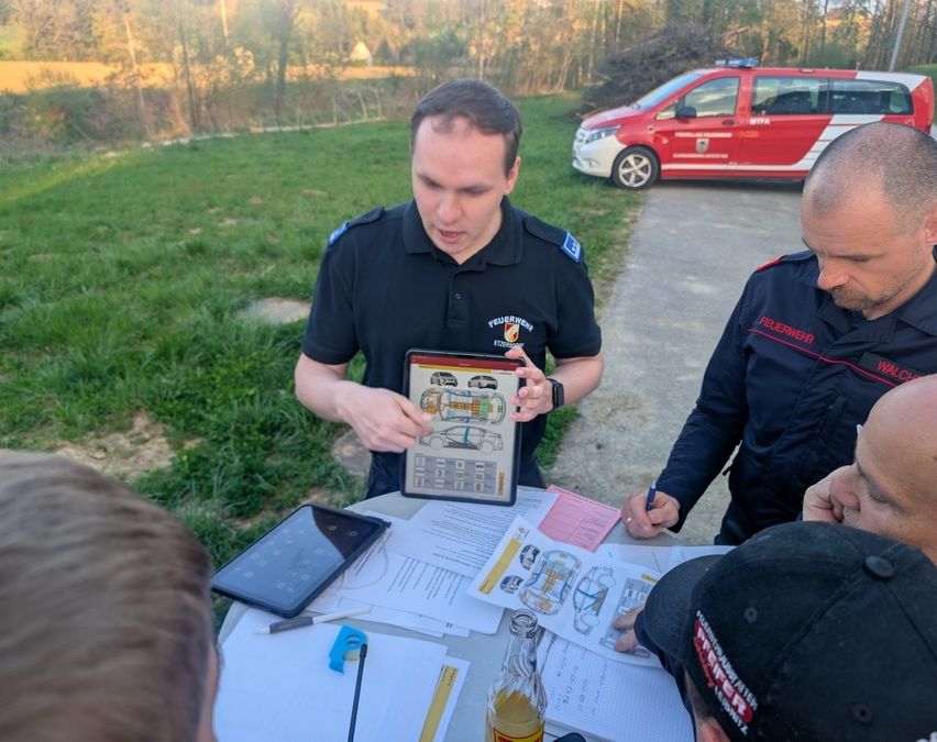 Ein Feuerwehrmann überprüft ein Bild auf einem Tablet, während ein anderer Feuerwehrmann Notizen macht. Dahinter ist ein Feuerwehrwagen geparkt.