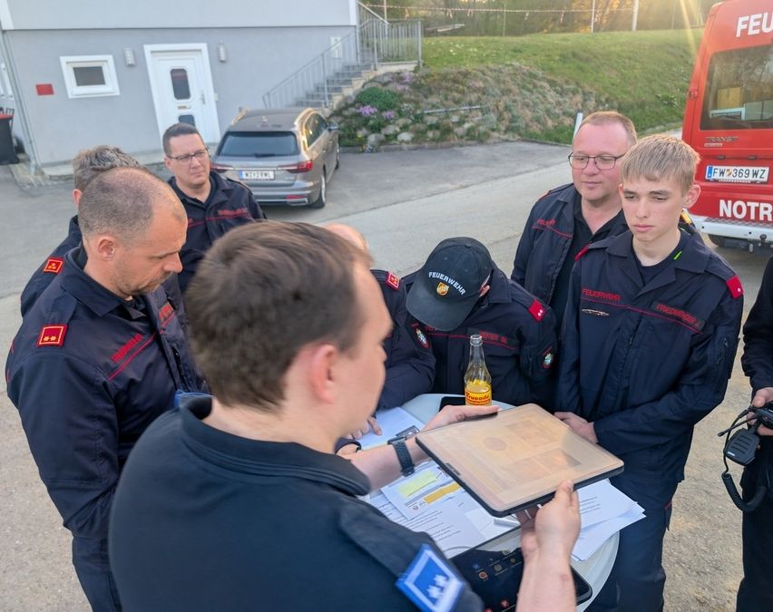 Eine Gruppe von Feuerwehrleuten in Uniform steht um einen Tisch mit einer Flasche und Papieren herum und bespricht möglicherweise einen Plan.
