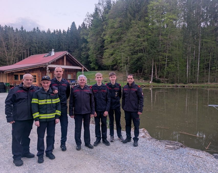 Eine Gruppe Feuerwehrleute in Uniform posiert für ein Foto vor einem kleinen See mit einem Holzhaus im Hintergrund.
