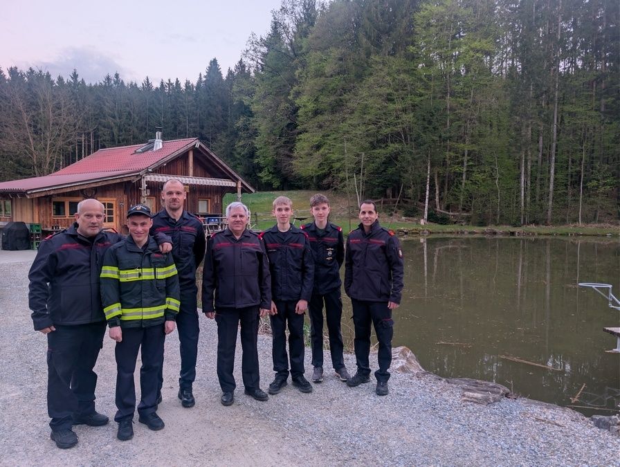 Eine Gruppe Feuerwehrleute in Uniform posiert für ein Foto vor einem kleinen See mit einem Holzhaus im Hintergrund.