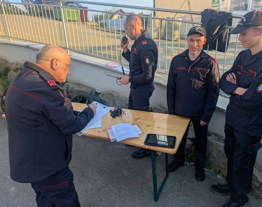 Drei Männer in Uniform stehen um einen Tisch herum mit Papieren, einem Tablet und einem Funkgerät. Ein Mann schreibt auf einen Clipboard, während ein anderer ein Funkgerät hält. Ein dritter Mann schaut zu.