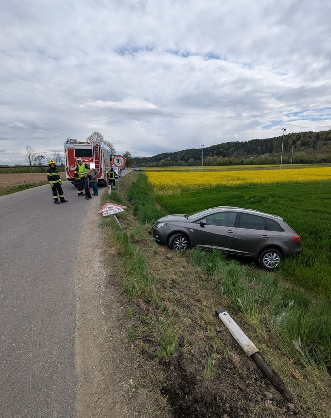 Ein Autounfall ereignete sich auf einer Straße. Ein graues Auto ist von der Straße abgekommen, mit Rettungskräften am Unfallort, einschließlich Feuerwehrleuten und Rettungskräften. Ein Feuerwehrwagen ist in der Nähe geparkt.