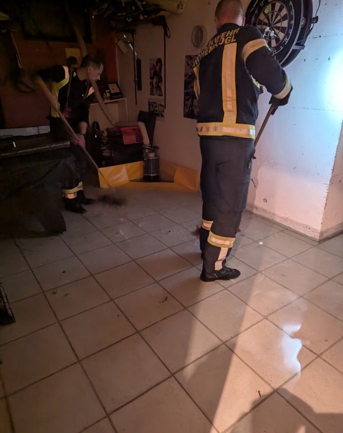 Zwei Feuerwehrleute säubern einen Raum mit gefliesten Böden, Besen und einem Eimer.