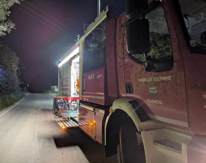Ein Feuerwehrwagen aus Hohenkogl steht bei Nacht mit offener Tür und eingeschalteten Lichtern am Straßenrand.