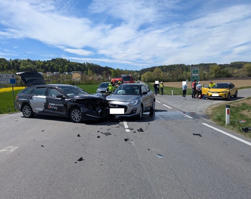 Ein Autounfall auf einer Straße mit zwei beschädigten Autos und Menschen darum herum. Bäume und ein Gebäude sind im Hintergrund.