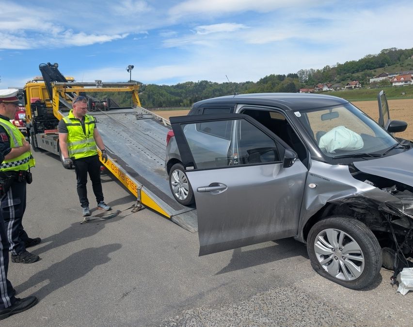 Ein Abschleppwagen zieht ein beschädigtes graues Auto auf einer Straße. Zwei Männer in Warnwesten stehen in der Nähe.