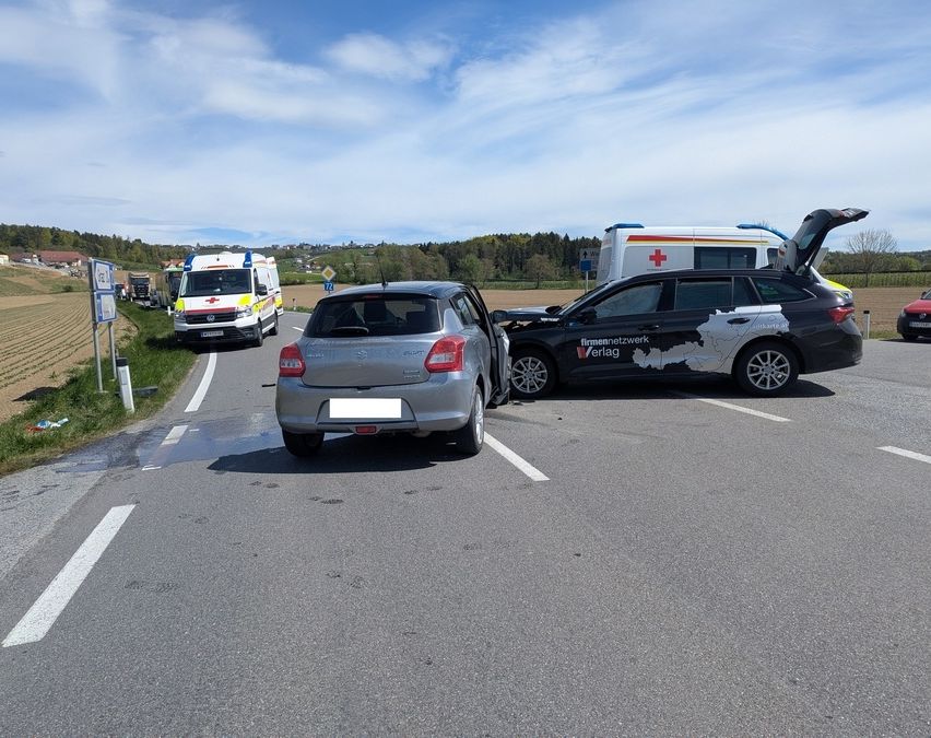 Ein Autounfall auf einer Landstraße mit anwesender Ambulanz und Feuerwehr. Ein schwarzes Auto und ein silbernes Auto sind beteiligt.
