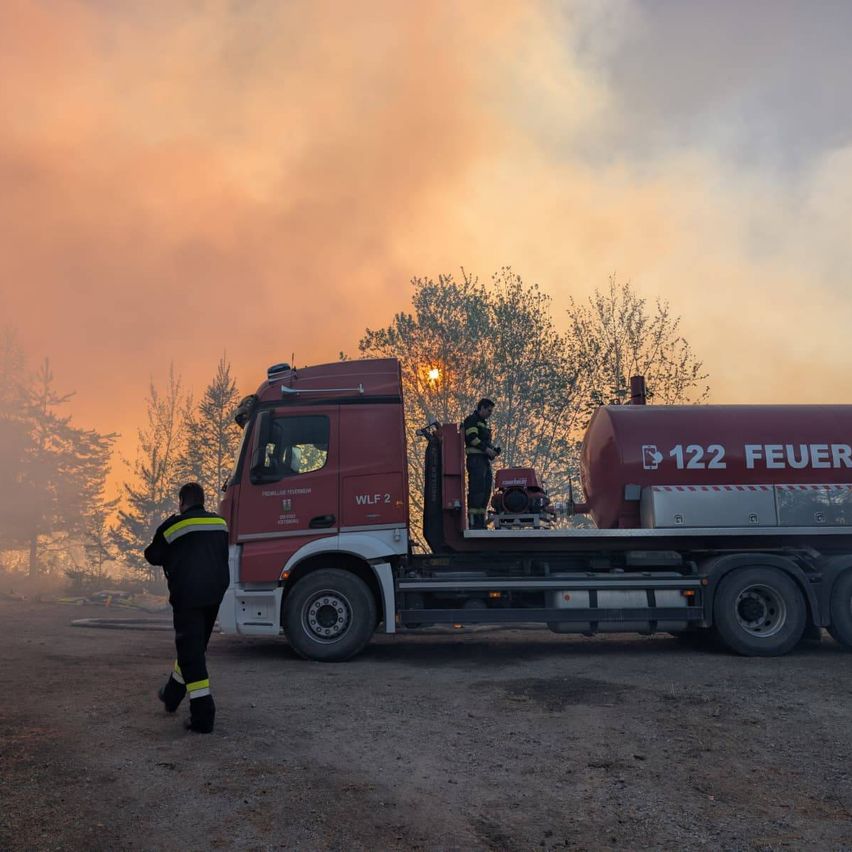 Ein Feuerwehrmann geht an einem roten Feuerwehrauto vorbei, ein anderer steht auf dem Auto. Es gibt Bäume und einen rauchigen Himmel im Hintergrund.