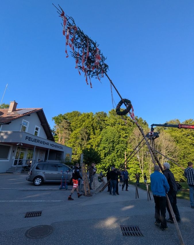 Menschen versammeln sich um einen hohen Pfahl mit Bändern vor dem Feuerwehr Hohenkogl Gebäude. Ein Kran ist in der Nähe positioniert, und ein Auto ist am Eingang geparkt.