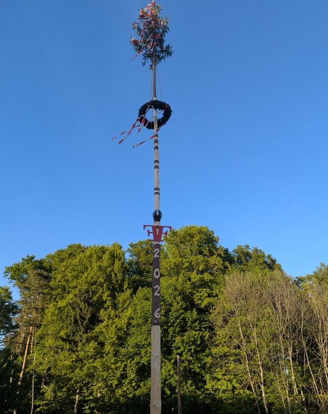 Ein hoher Mast mit der Aufschrift 2026, gekrönt von einem Kranz und umgeben von Bäumen.