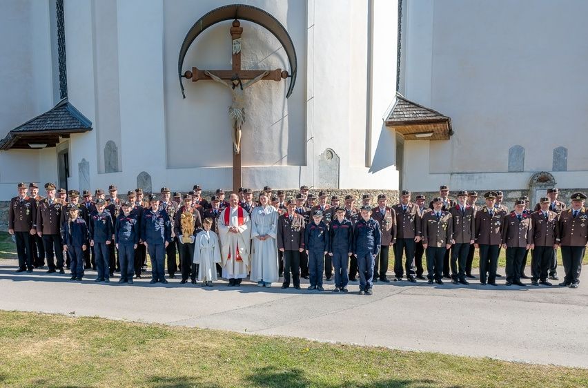 Eine Gruppe uniformierter Personen steht vor einer Kirche mit einem großen Kreuz an der Wand. Ein Priester und eine Frau stehen in der Mitte.