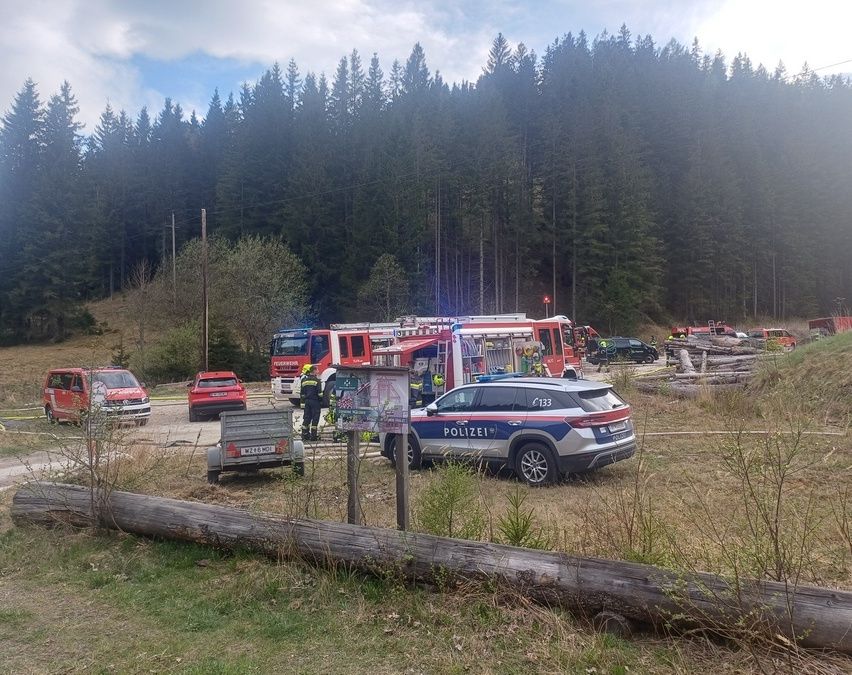 Einsatzfahrzeuge, darunter ein Polizei-SUV, sind auf einer unbefestigten Straße mit bewaldetem Hintergrund geparkt. Eine Person mit gelbem Helm steht neben einem Feuerwehrwagen.