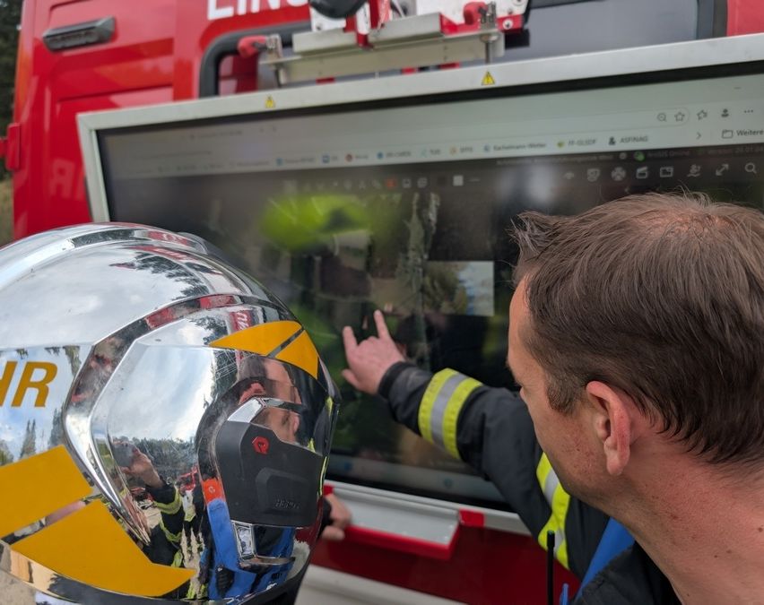 Ein Feuerwehrmann mit einem reflektierenden Helm zeigt auf einen großen Monitor, wahrscheinlich um Vorfallaufnahmen zu überprüfen. Der Monitor zeigt ein Bild eines Feuers. Der Ort ist draußen, in der Nähe eines Feuerwehrwagens.