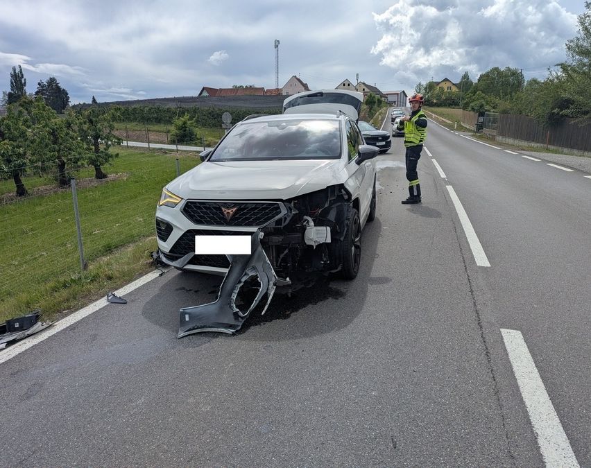 Ein weißer Wagen mit beschädigter Front steht am Straßenrand. Eine Person in einer reflektierenden Weste steht in der Nähe.