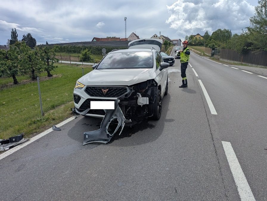 A white car with a damaged front is parked on the side of the road. A person in a reflective vest is standing nearby.