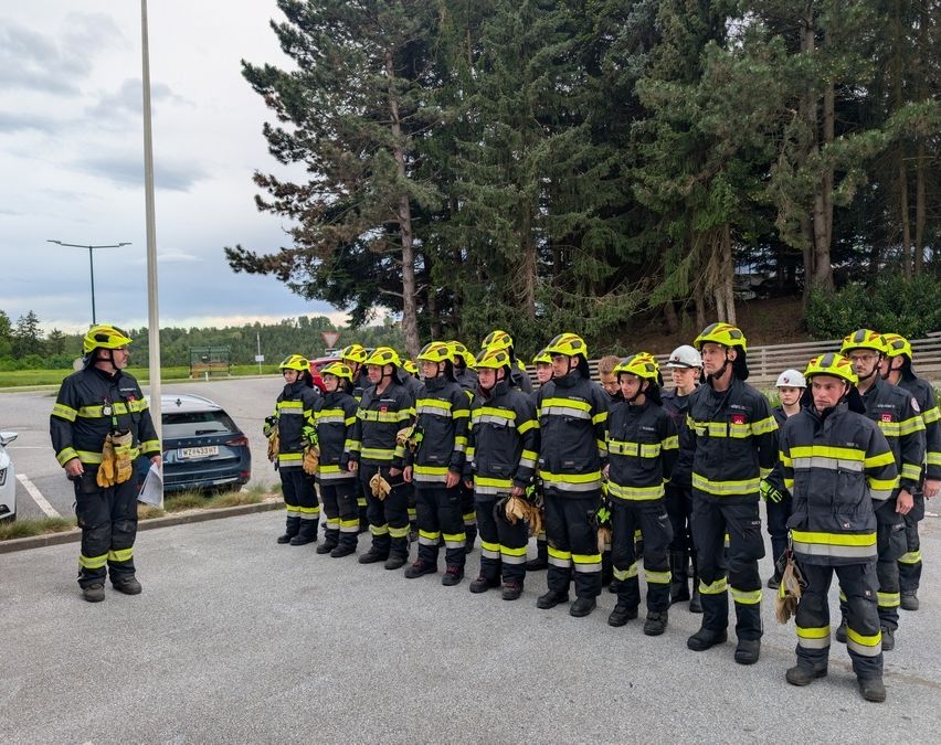 Eine Gruppe von Feuerwehrleuten in Uniform steht in einer Reihe auf einem Parkplatz mit Bäumen und einem Auto im Hintergrund.