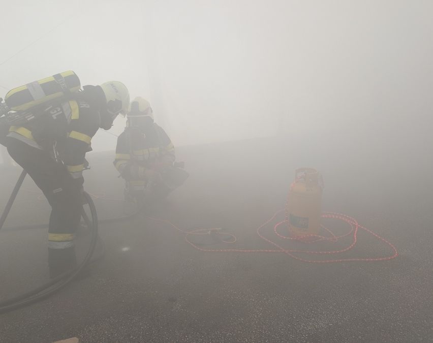 Zwei Feuerwehrleute in Schutzkleidung arbeiten in einer nebligen Umgebung, einer hält einen Schlauch und der andere einen Gaszylinder.