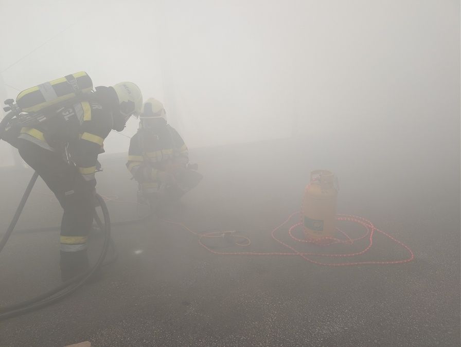 Zwei Feuerwehrleute in Schutzkleidung arbeiten in einer nebligen Umgebung, einer hält einen Schlauch und der andere einen Gaszylinder.