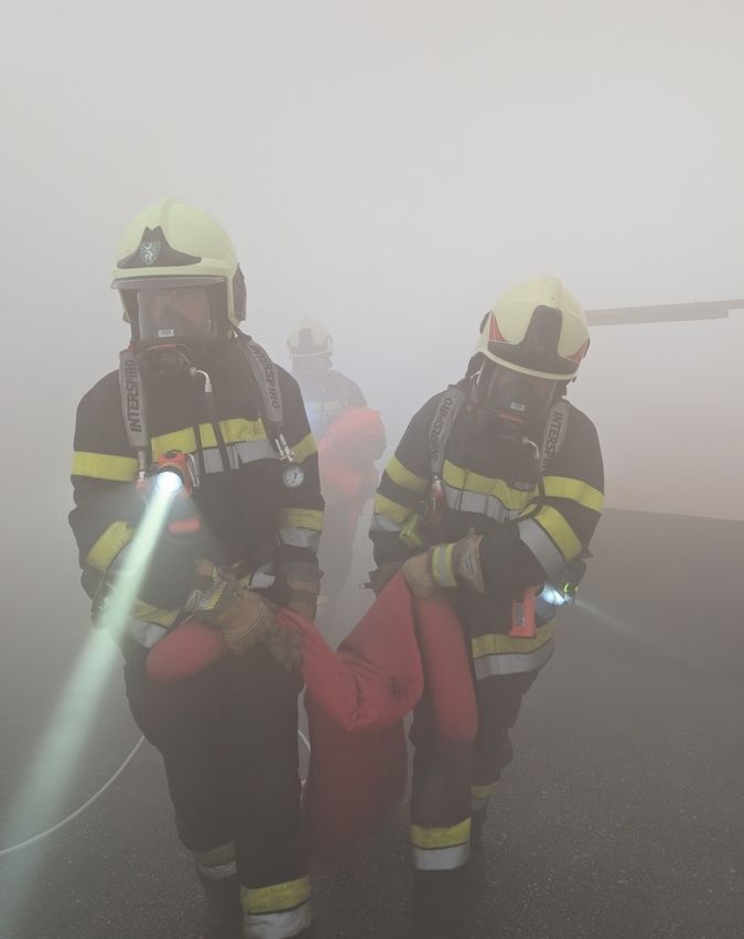 Zwei Feuerwehrleute tragen ein rotes Objekt, während sie Helme tragen und Taschenlampen halten. Sie befinden sich in einem nebligen Bereich, mit einem weiteren Feuerwehrmann im Hintergrund.